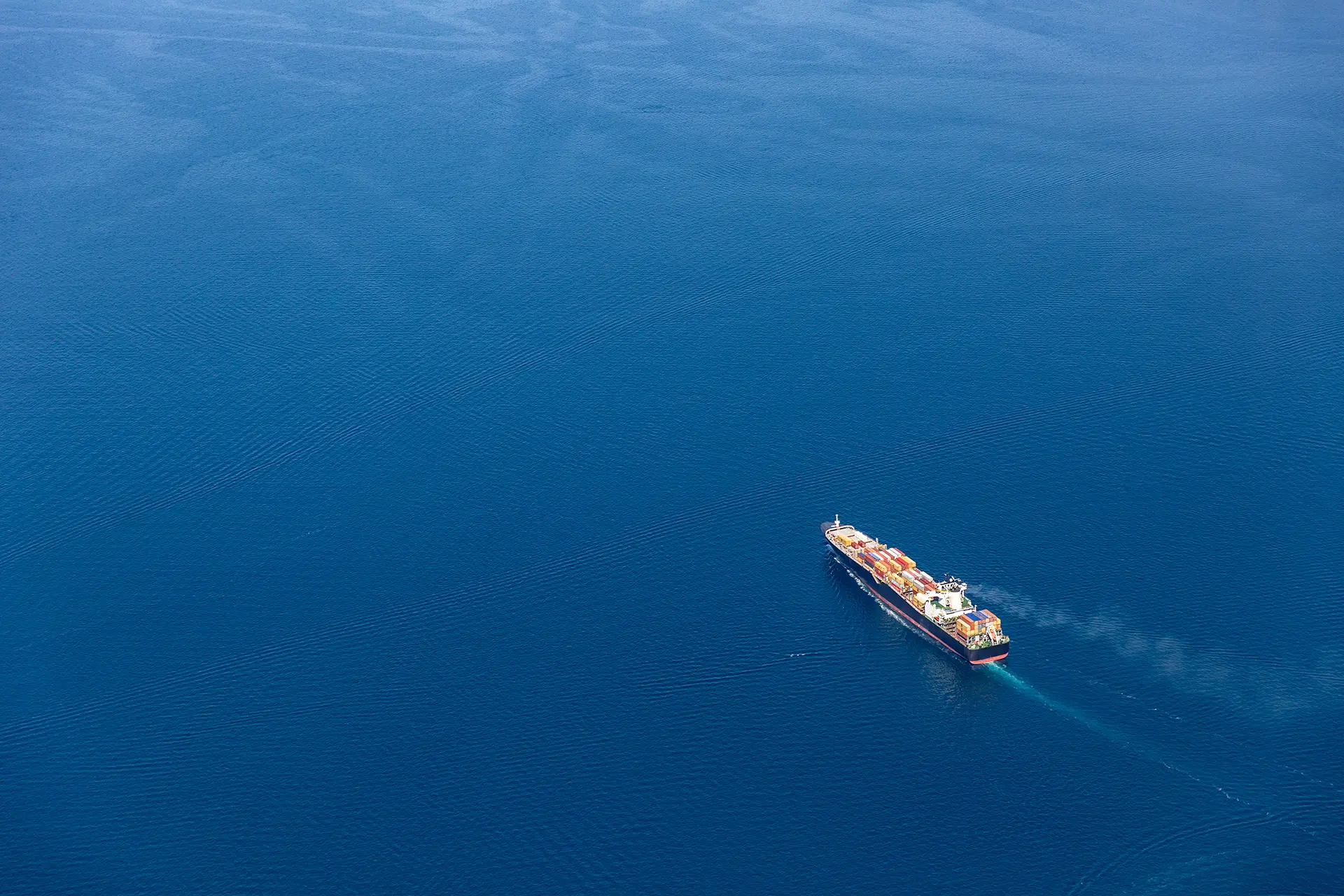 A cargo ship with colorful containers sails through calm blue ocean, seen from above in the bottom-right quadrant
